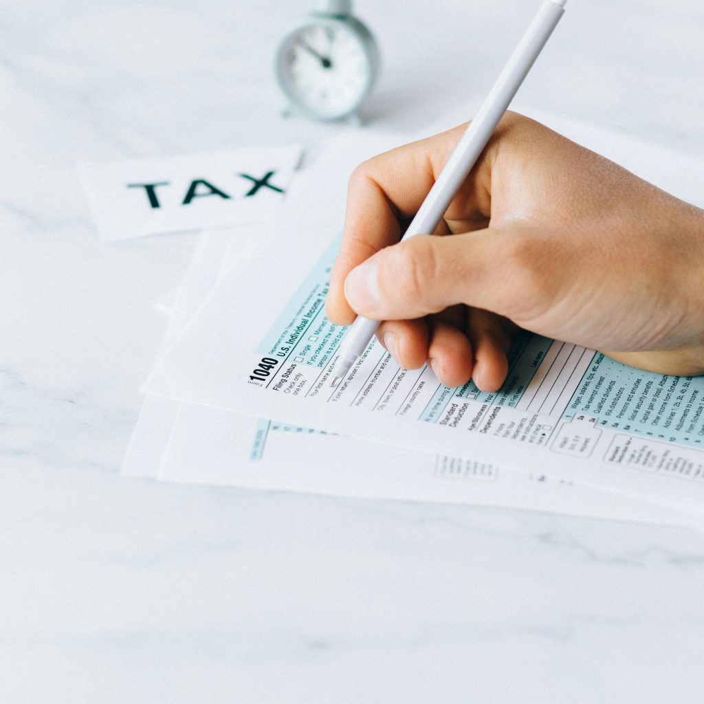 A person's hand holds a white pen, filling out a Form 1040 U.S. tax return on a bright, marble surface with a small alarm clock and a "TAX" label in the background.