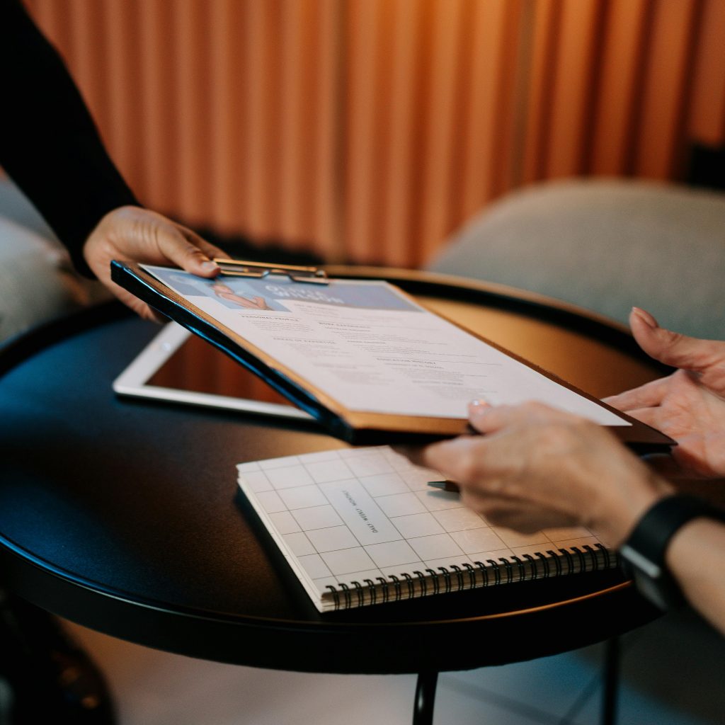 Two people's hands exchange a resume or document on a clipboard across a small round table during an interview or meeting.