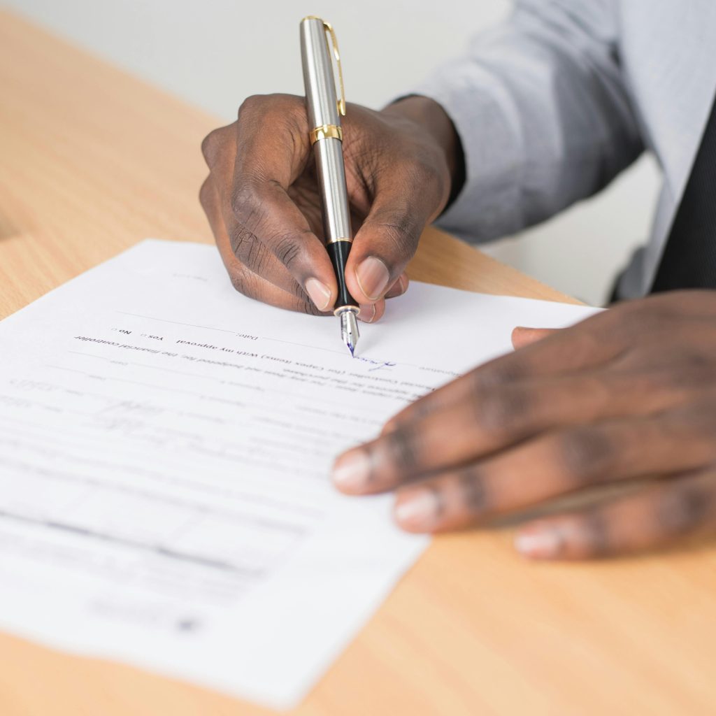 A close-up of a person's dark skin hands signing a paper document on a light wooden desk with a silver and gold pen, wearing a suit and tie.