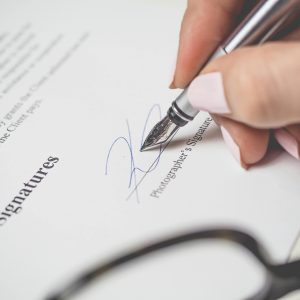  A hand with light pink nail polish signs a document, labeled "Signatures," with a fountain pen, next to a blurred pair of glasses.