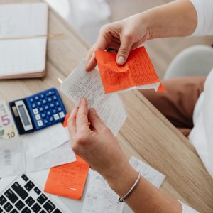 A person's hands sort through a pile of paper receipts and orange receipts on a wooden desk next to a calculator and laptop keyboard.