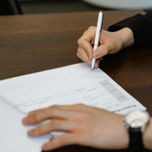  A person's hands sign or write on an open document or report with a white pen at a dark wooden desk; a watch is visible on the left wrist.