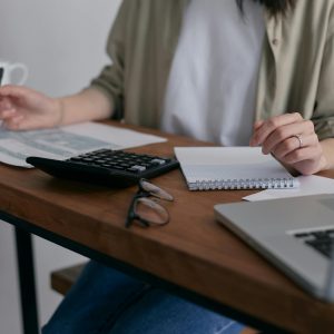  A person sits at a wooden desk reviewing tax forms and holding a notebook, with a calculator, laptop, glasses, and coffee mugs nearby.