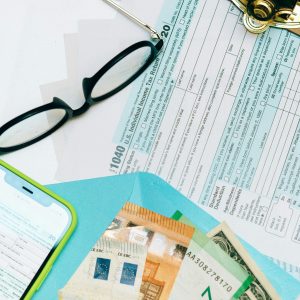  An overhead view of a US 1040 Income Tax Return form next to reading glasses, Euro currency, and a smartphone displaying a tax app.