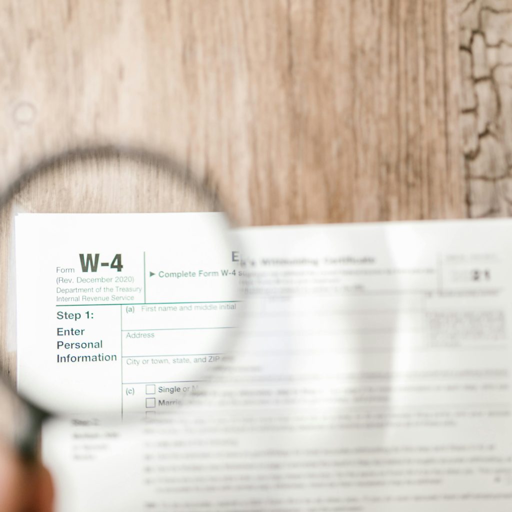 A magnifying glass held over an IRS Form W-4 (Employee's Withholding Certificate), making the text on a wooden desk clear.