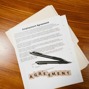  An Employment Agreement document with a pen on it, resting on a wooden surface, with Scrabble tiles spelling out AGREEMENT.